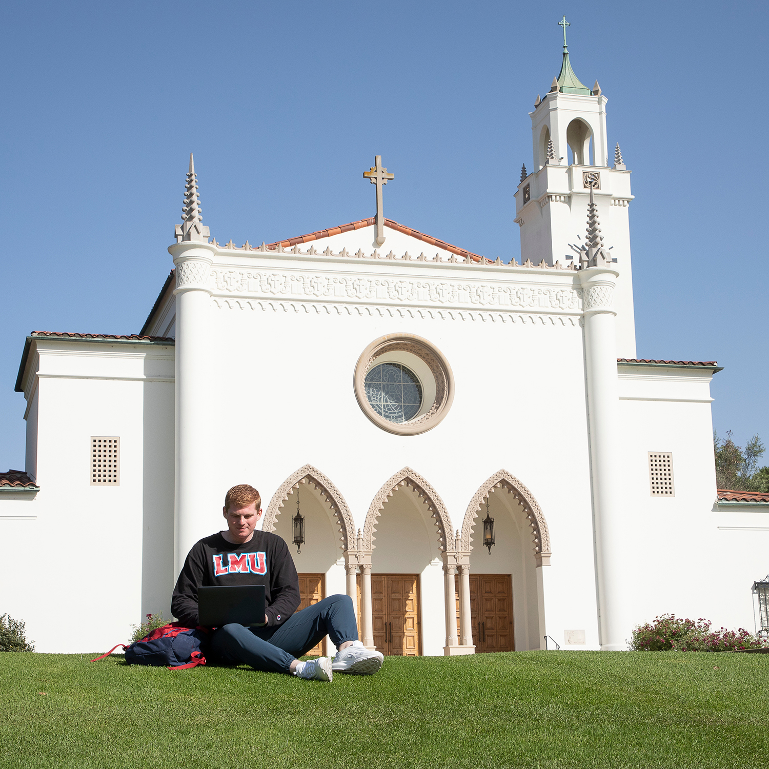man in LMU sweater sitting on the lawn in front of Sacred Heart Chapel